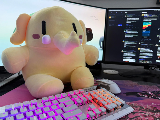 A photo I took of the plush Mastodon stuffed animal that arrived. #Plushtodon is the hashtag used for these photos and it's even on the tag on the ear. 

You can see part of my keyboard and two monitors in the photo along with my mouse.