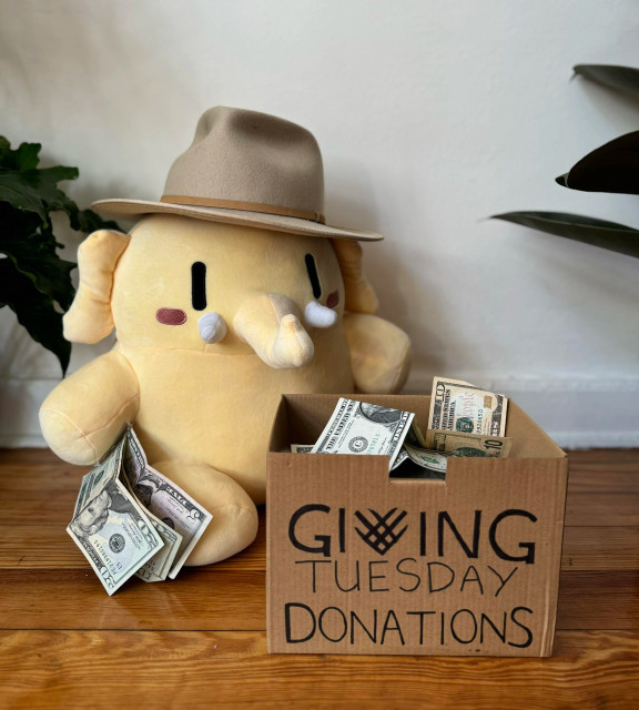 A cute stuffed Mastodon toy wearing a hat, sitting in front of a box labelled "Giving Tuesday donations" filled with US dollar bills.