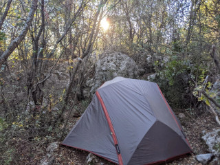 A tent in the forest surrounded by rocks, sun going through the branches.