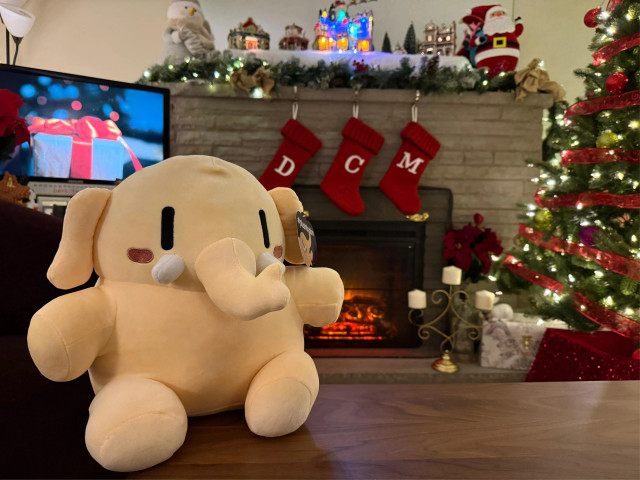 A yellow plush mastodon sitting in a room of Christmas decorations. There’s a tree to the right, a fireplace with stockings behind, a village scene on the mantel above, and a screen showing a picture of a present to the left. 