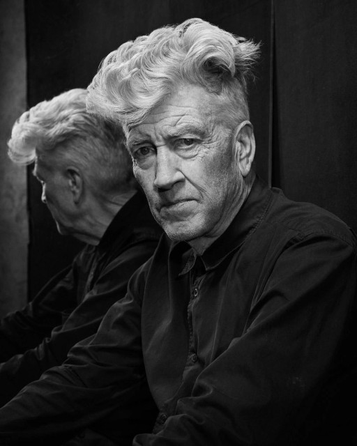 Black-and-white photograph of the director David Lynch, an elderly white man with a shock of white curly hair. Lynch is wearing a dark button-down shirt, and sitting in half profile in front of a mirror. The back of his head is partially visible behind him, creating an eerie double effect.