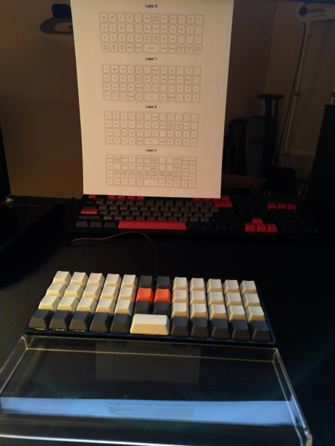 a 40% ortholinear keyboard in the foreground, a cheat sheet describing all the layers above it, a full-sized keyboard in the background