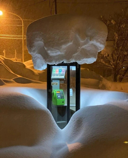 Photography.  A color photo of a snowy landscape with an illuminated telephone booth in the dark. The snow in the photo forms round hills and is over a meter high. In the middle of these white hills is an illuminated glass telephone box with a green telephone set. Someone probably cleared the snow the day before, because there is a pile of snow twice as wide and about 1.50 meters high on top of the phone booth. In the background, another light source illuminates the white surroundings and makes them appear orange. A strange atmosphere between cold and warmth, with round snow shapes and a telephone box that seems to have landed in it like a UFO. It's a harsh winter on the island of Hokkaido, but at least ... you can make a phone call.