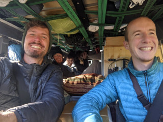 A few people in a very small van-style car, everybody is smiling, a lot of tools and different things are around, stabilized with the straps and attached to roof or sides.