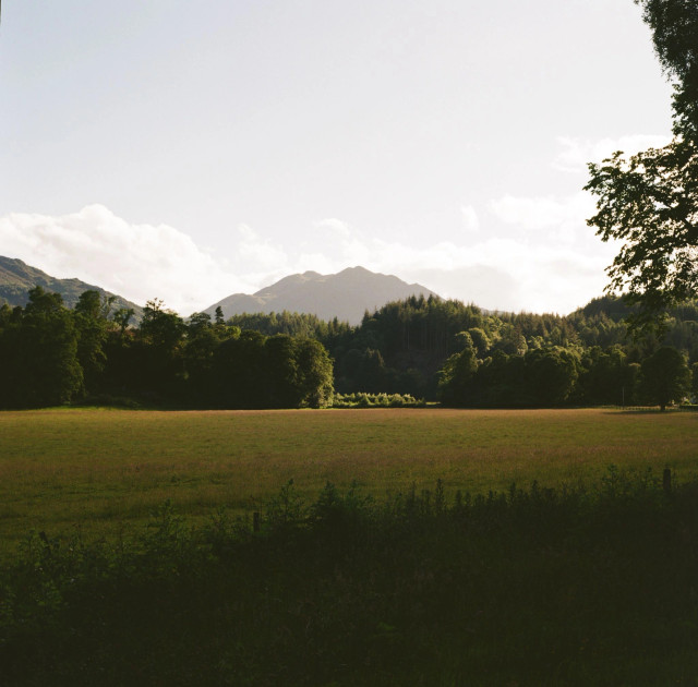 A colour photograph of a late evening scene on summer solstice looking over fields, trees and a mountain 