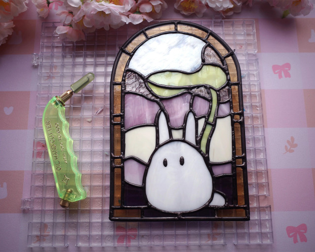 Stained glass art featuring Chibi Totoro in yellow, pink, and purple theme. Shown in workspace with glass cutter tool and framed with pink blossoms.