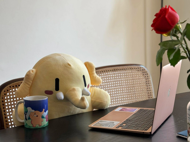 A cute stuffed Mastodon toy holding a Mastodon mug while sitting in front of a laptop.