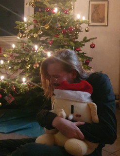 An image of Erik Uden and Oskar the Mastodon plushie snuggling in front of a Christmas tree. In the background is an image of Friedrich Uden, the oldest surviving visual record of a 19th century ancestor of Erik Uden.