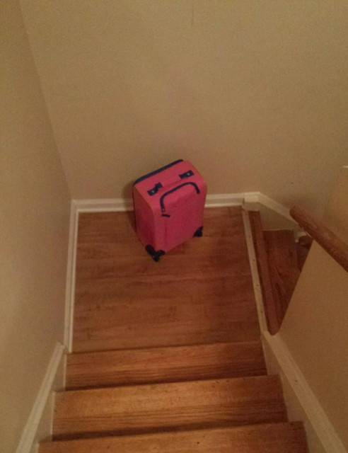 A photograph of a pink suitcase on a wooden staircase. It's handle is an upside down U shape, like a grimace. The attachments on top like upset eyes glaring.