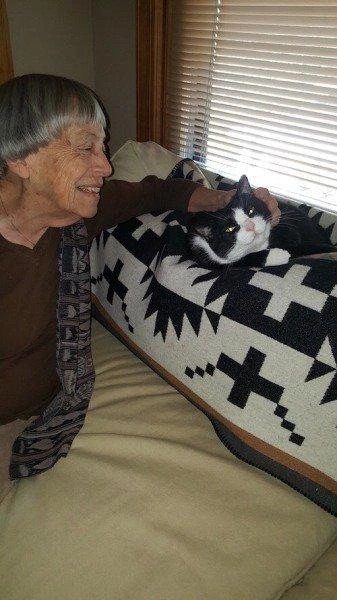 Ursula K Leguin sitting on a sofa, with her tuxedo cat curled up near her, as she is lovingly leaning forward and petting him, the cat's head is slightly turned towards her and he clearly looks content.