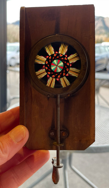 fair skinned hand holding up a wooden kaleidescope in front of a window. rectangular piece of wood with a intricately colored circular piece of glass inserted in the middle and a rod and small handle are attached to the lowest part of the circle of glass